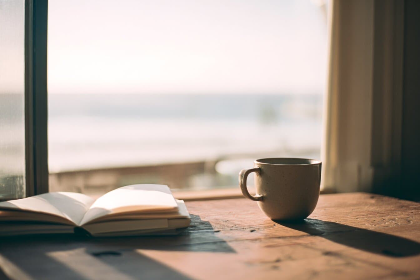 Luce del mattino su una scrivania di legno con un quaderno e una tazza, accanto a una finestra che dà sul mare.
